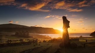 one moai statue in the foreground has beams of sunlight radiating from behind and there is a row of moai statues in the background.