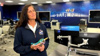 a smiling woman holds a small plush narwhal toy in a room with desks and computer monitors