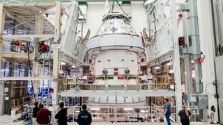 a cone-shaped spacecraft surrounded by scaffolding in a big warehouse. people stand in front looking at it