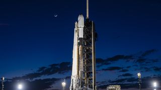 A white and black SpaceX rocket with the Intuitive Machines IM-1 moon lander aboard on the launch pad with a crescent moon overhead.