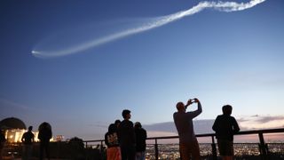 an expanding white plume extends from a small point of white light on the left, growing thicker in a stream across the darkening blue sky. Silhouettes of onlookers line the bottom, near a guard rail fence. City lights are seen low in the distance.