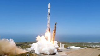 a black and white spacex falcon 9 rocket launches into a blue sky with the ocean in the background