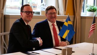 Two men in dark suits shake hands while sitting at a table with documents and two small flags. Both men are wearing glasses.