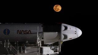 the top portion of a white rocket is seen lying on structural supports in the black of night. An unseen light source shines down the rocket from the nose. An orange/yellow moon hangs in the sky above.