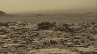 A dull yellow-tinted, rocky terrain blown with dust, as the sky in the distance matches the ground's dusty tones.