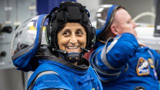 two astronauts in spacesuits with visors open. the woman astronaut in front turns to smile at the camera while the man astronaut in back looks to the side